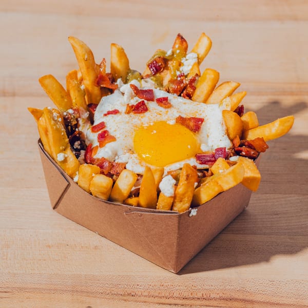 Loaded fries topped with a fried egg, bacon, cheese, and sauce in a cardboard serving tray on a wooden table.