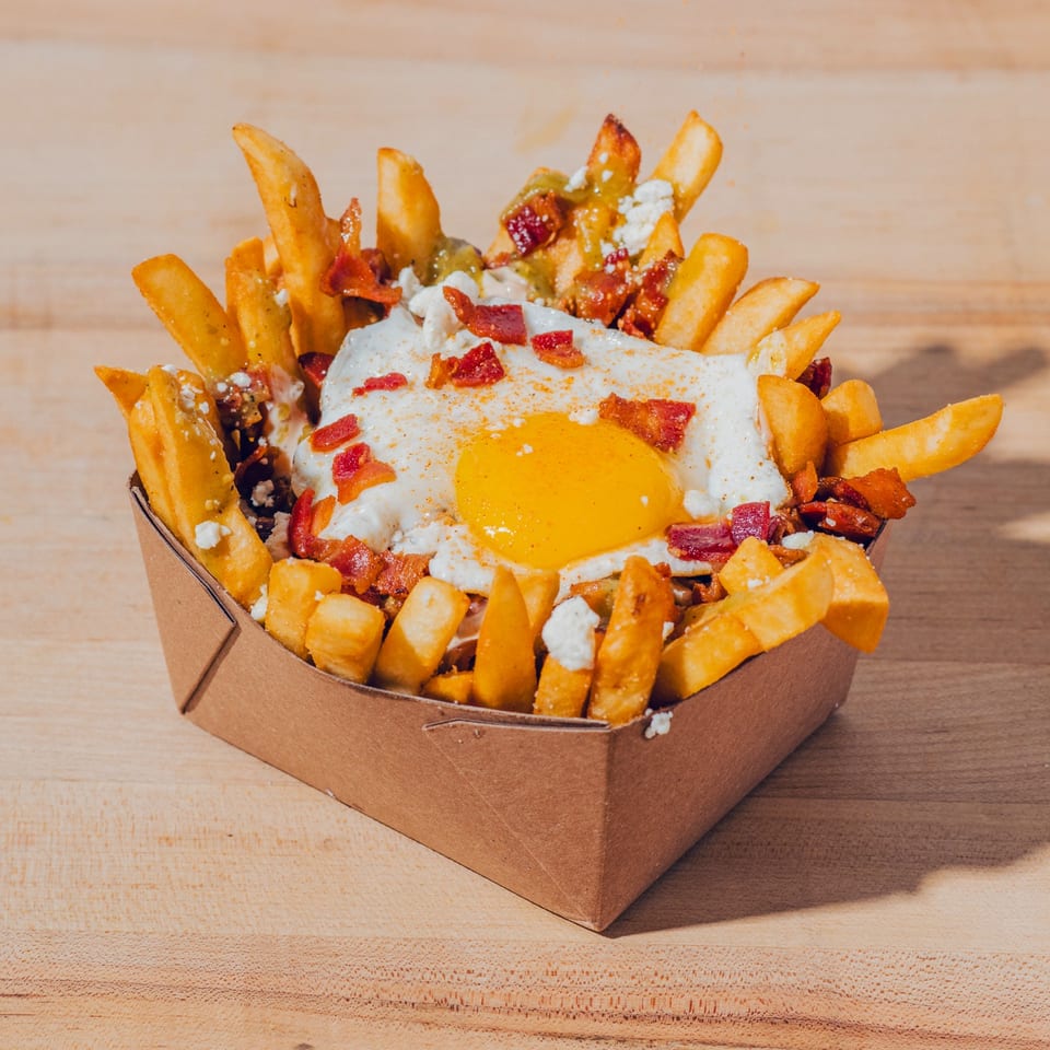 Loaded fries topped with a fried egg, bacon, cheese, and sauce in a cardboard serving tray on a wooden table.