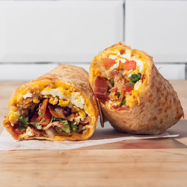 Two breakfast burrito halves filled with scrambled eggs, melted cheese, bacon, tomatoes, mushrooms, and herbs on a wooden table.