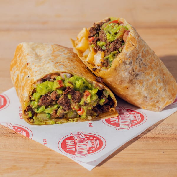 Two burritos stuffed with seasoned meat, guacamole, vegetables, and fries sit on branded paper atop a wooden table.