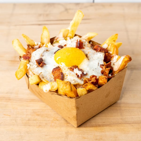 Fries topped with a fried egg, bacon bits, and cheese in a cardboard container on a wooden surface.