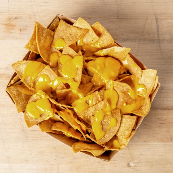 Crispy tortilla chips generously covered in melted yellow cheese sauce, served in a cardboard tray.