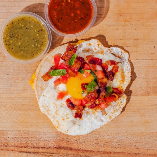 Overhead view of a fried egg topped with bacon, tomatoes, peppers, cilantro, and cheese on a tortilla, served with green and red salsa.