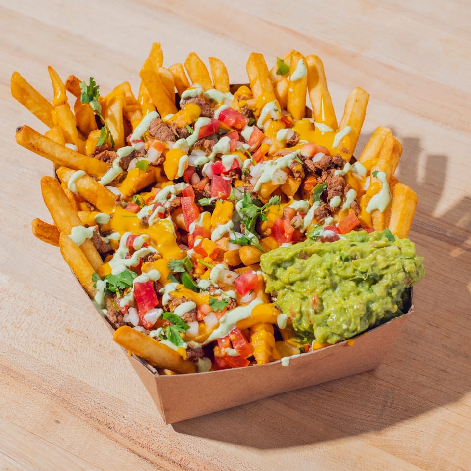 Loaded fries topped with seasoned meat, cheese sauce, tomatoes, cilantro, creamy green sauce, and guacamole in a paper tray.